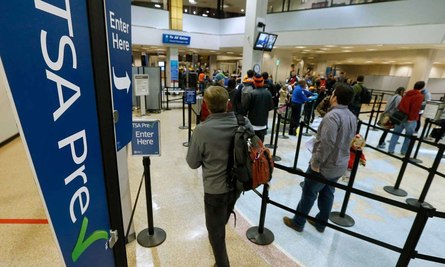 Traveler with backpack waits in line at an airport security checkpoint, surrounded by tall blue 'Enter Here' signs.
