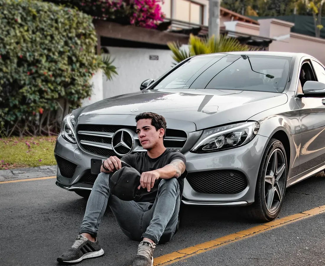 Young man sits on the street beside a silver Mercedes sedan in a residential area.