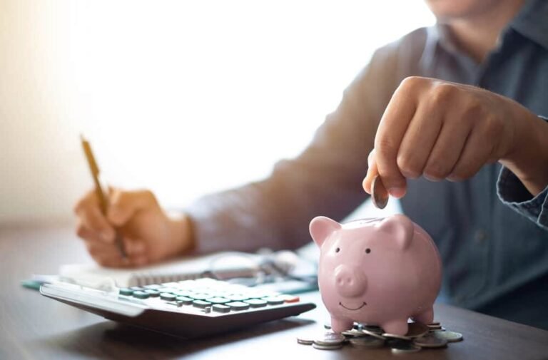 Person places coins into a pink piggy bank while using a calculator and writing with a pencil for budgeting.