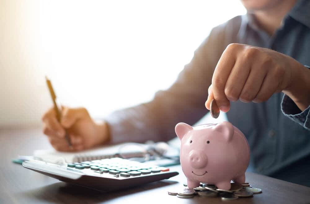 Person places coins into a pink piggy bank while using a calculator and writing with a pencil for budgeting.