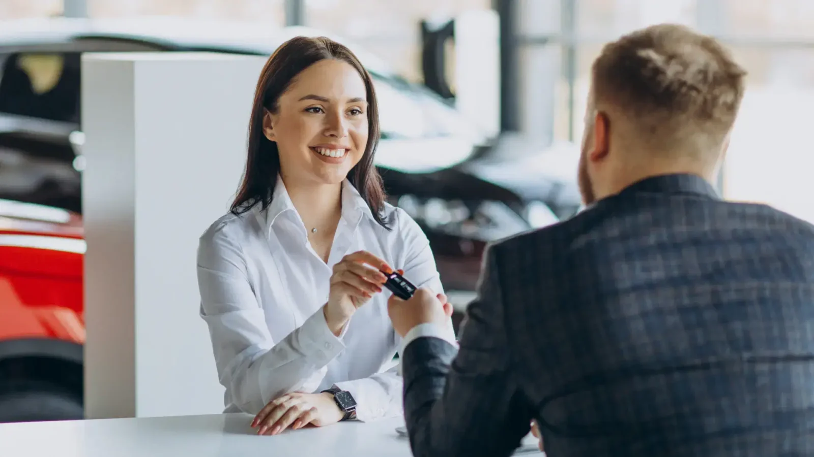 Saleswoman in a white shirt hands car keys to a customer at a dealership, smiling across a desk in a showroom.