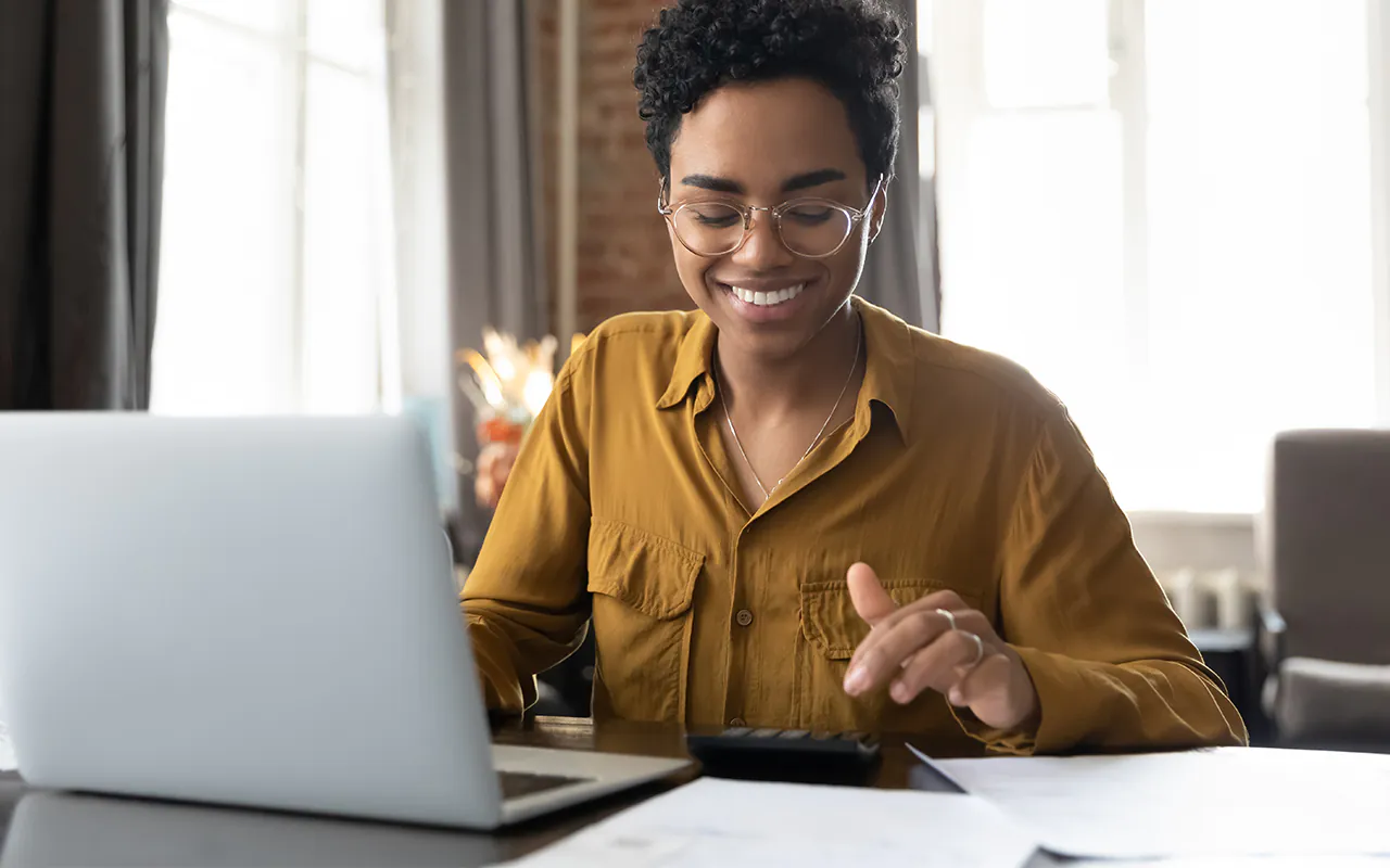 connexus-credit-union-cd-review | Beelinger Person with short curly hair and round glasses smiles while using a silver laptop at a desk with papers nearby in a bright room.