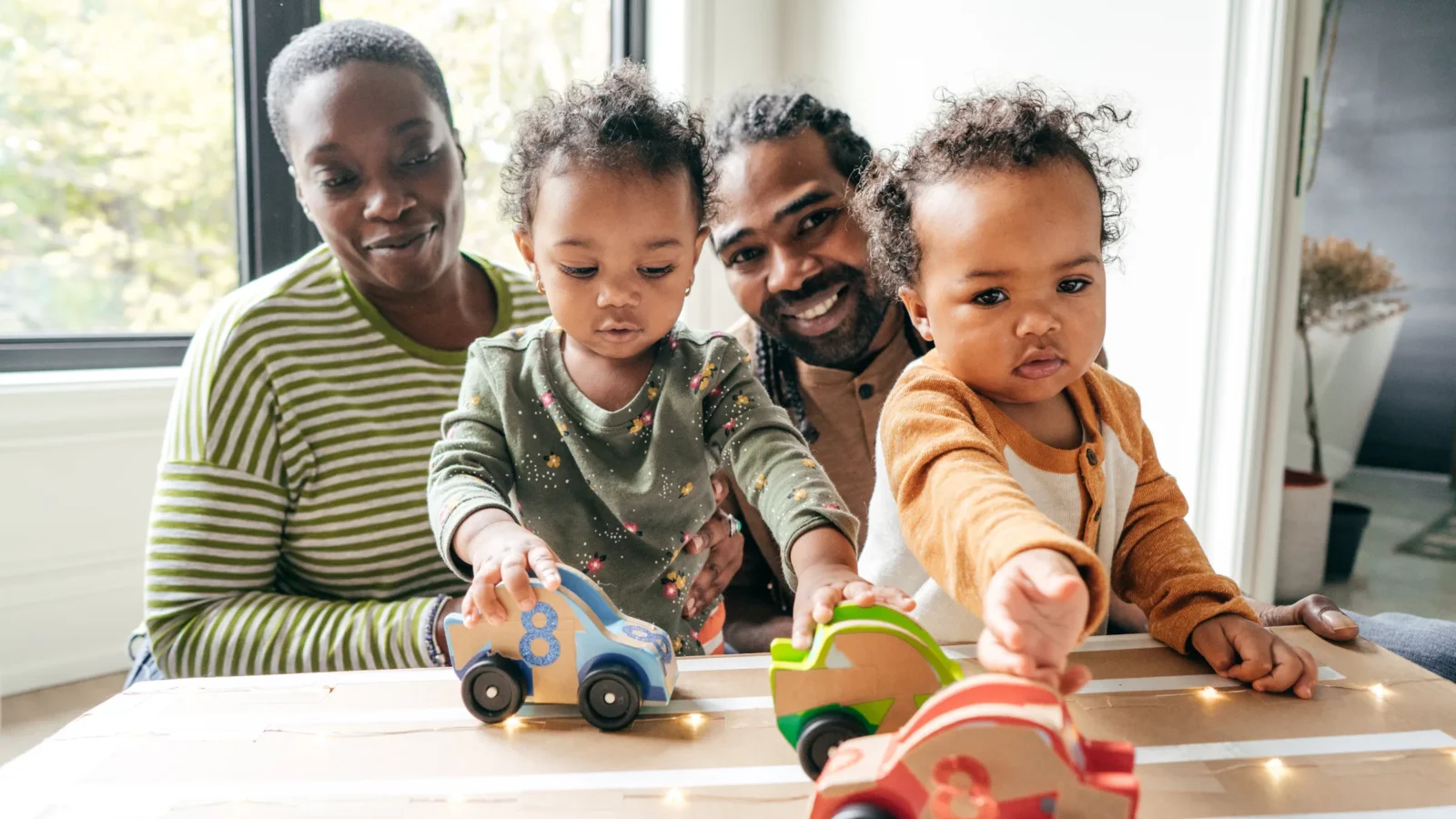 Family of four playing with colorful wooden toy cars on a track indoors by a window.