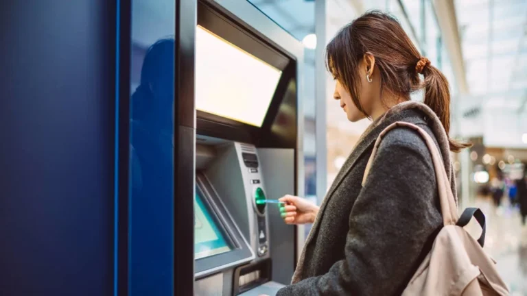 Woman inserting a card into an ATM in a bright indoor setting, with a backpack on her shoulder