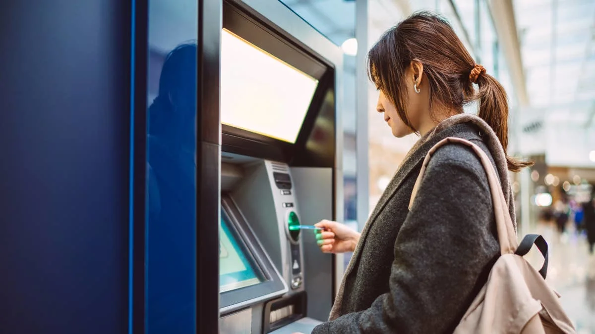 Woman inserting a card into an ATM in a bright indoor setting, with a backpack on her shoulder