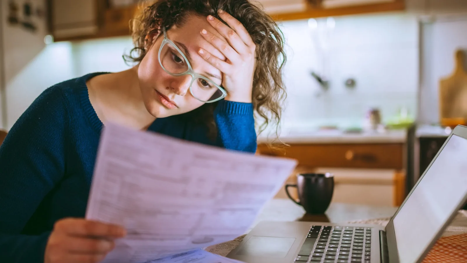Woman with glasses sitting at a laptop, holding a paper and rubbing her forehead in a kitchen, appearing stressed or overwhelmed by bills.